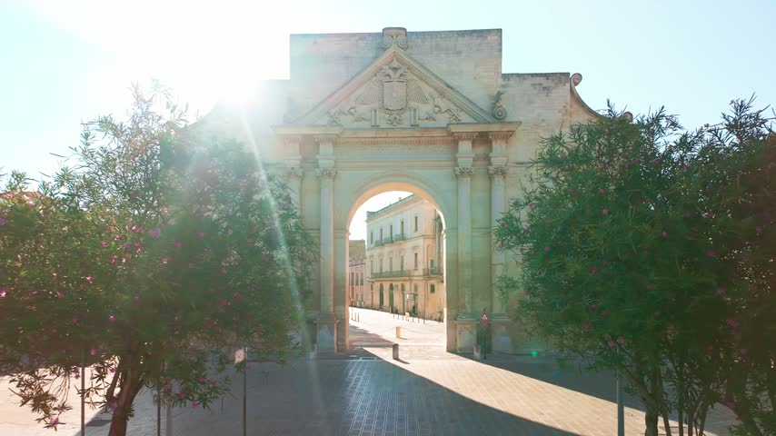 The historic Porta Napoli triumphal arch framed by green trees on a sunny day in Lecce, Apulia, Italy.