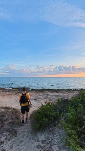 Vertical slow motion footage of a man walking from a sandy path to the shoreline at sunset, then stopping at the water