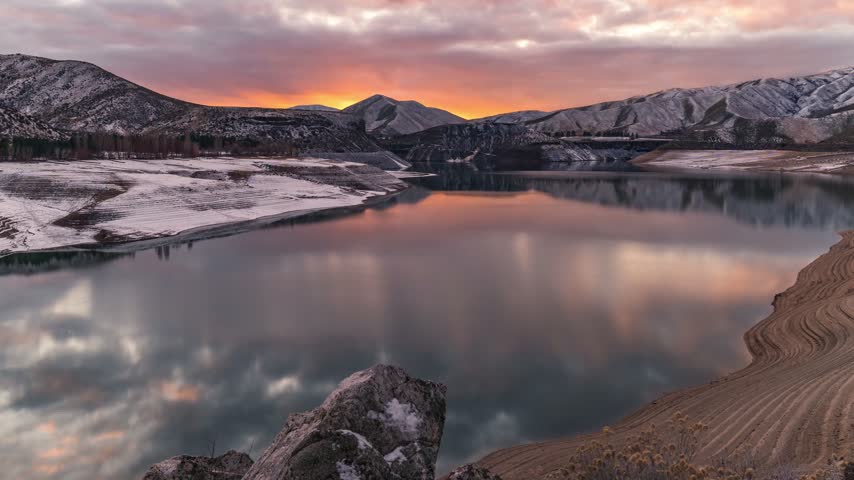 Lucky Peak Reservoir at sunset with a time lapse capturing vibrant orange clouds and snowy mountains reflecting on the still cold water during a serene winter evening