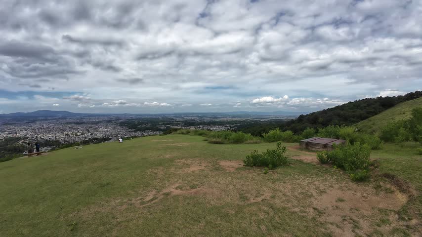 Wide hilltop summit view overlooking an expansive urban valley landscape under dynamic clouds. 