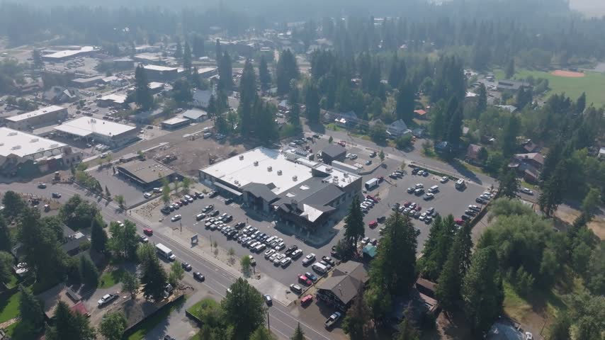 Aerial view showcasing a small town commercial hub with active vehicle traffic and parking lots surrounded by dense pine forests under a bright hazy sky