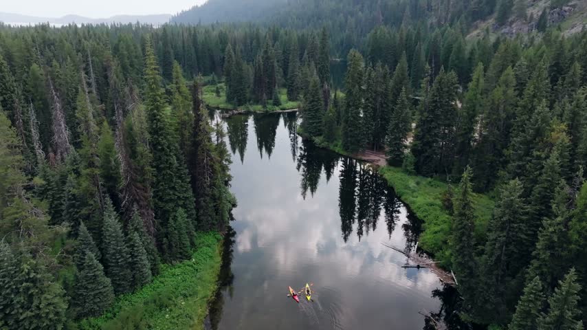 Aerial view of two kayakers paddling on a peaceful glass like mountain lake surrounded by dense evergreen pine forests in the wilderness of Idaho