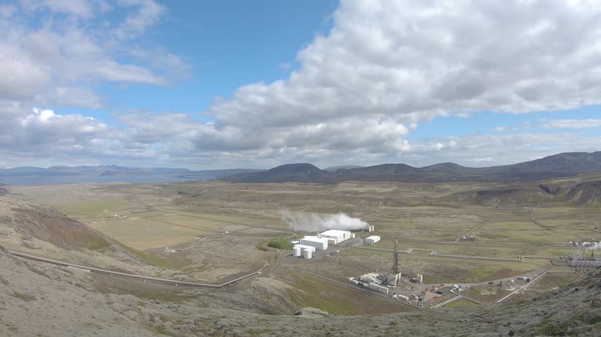 Nesjavellir, Iceland - 12 June 2019: Nesjavellir geothermal power plant releasing steam in a wide volcanic valley, with pipelines crossing the green valley floor.