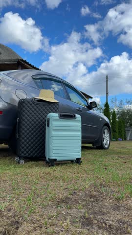 Luggage bags and a straw hat are positioned next to a parked car on grass, with a bright blue sky and clouds overhead, indicating travel plans