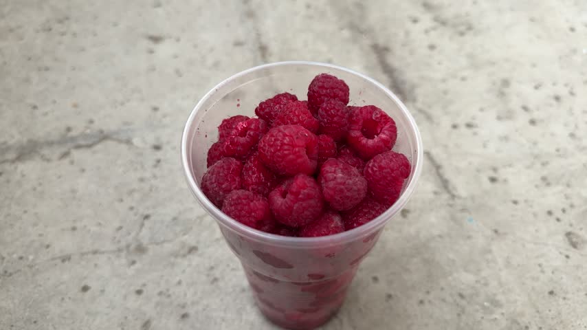 Fresh red raspberries in a clear plastic cup on a concrete surface outdoors, ripe berries, healthy snack, fresh fruit, summer harvest.