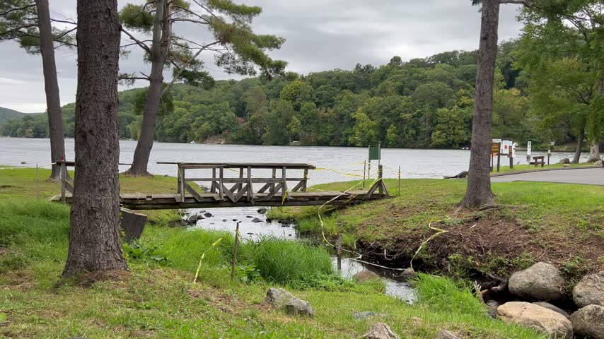 Small stream flows under a damaged wooden footbridge with caution tape blowing near a calm lake.