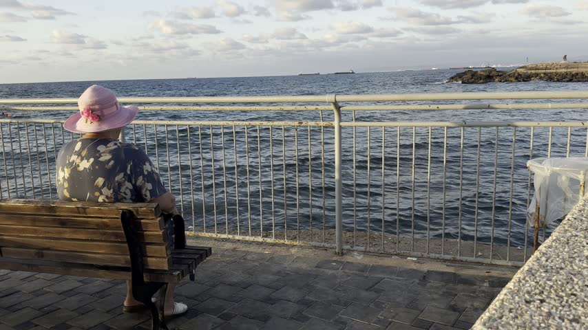 Haifa, Israel, 25 September 2026. An elderly woman in a colorful dress and wide-brimmed hat is sitting on a bench and looking at the sea, seen from behind.
