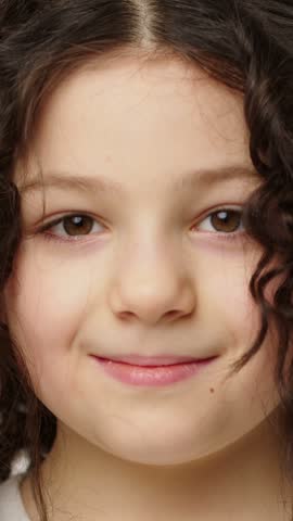 Close up portrait of a young girl with curly brown hair smiling toward the camera as her expression gradually becomes more open and cheerful