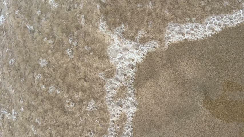 Close view of sea foam bubbles sliding across golden wet sand at the water line in Gandia, Spain.