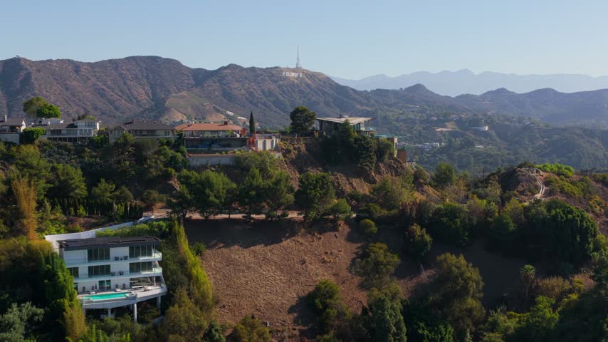 Cinematic aerial view of luxury hillside homes above a steep canyon in Los Angeles California. Modern villas with glass terraces and dry green ridges. Sunny peaceful day in Hollywood Hills.