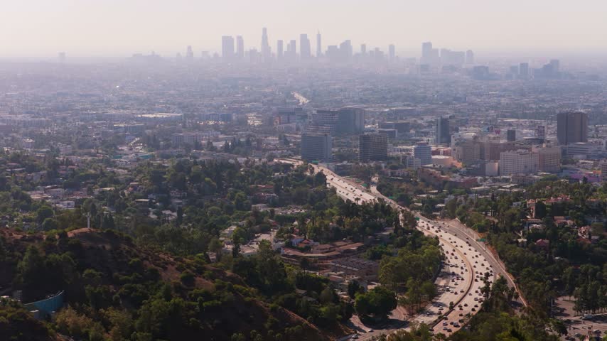 Cinematic aerial view of Los Angeles. Hazy downtown skyline on a hot summer day. Busy freeway traffic curving through Hollywood Hills. Dreamy California cityscape with smog and green hills.