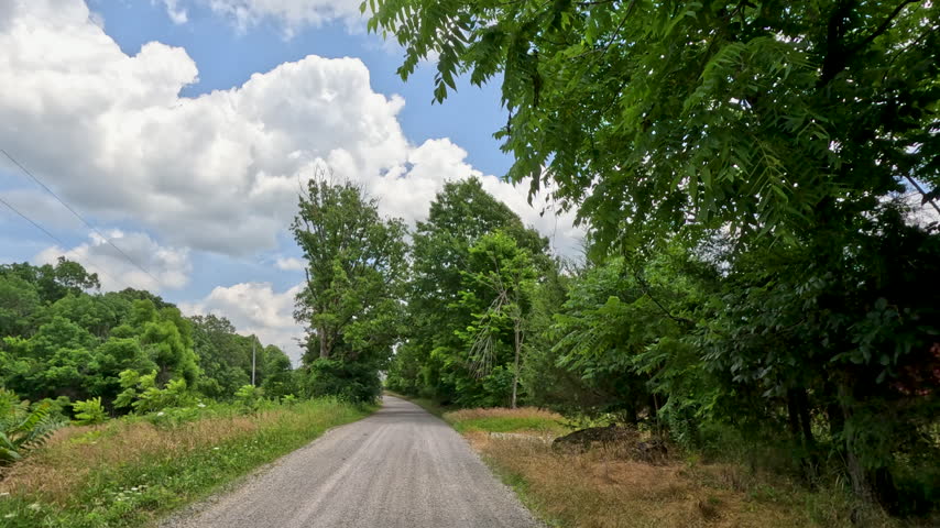Slow motion vehicle point of view shot starting from a stationary position and accelerating forward down a narrow, unpaved gravel country road surrounded by dense green forest and blue skies in rural Southern Illinois.