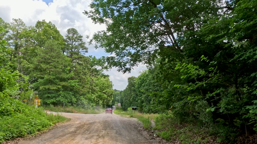 Slow motion vehicle point of view shot traveling behind another car that is kicking up a cloud of dust along a narrow, unpaved gravel country road surrounded by dense summer forest in Southern Illinois.