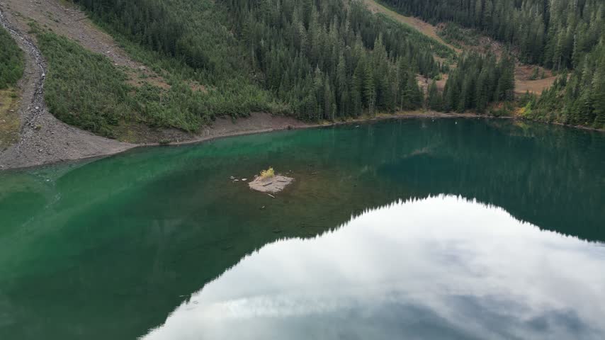 Still water in an alpine lake reflects passing clouds beneath a forested mountainside near Mount MacFarlane in British Columbia, Canada.