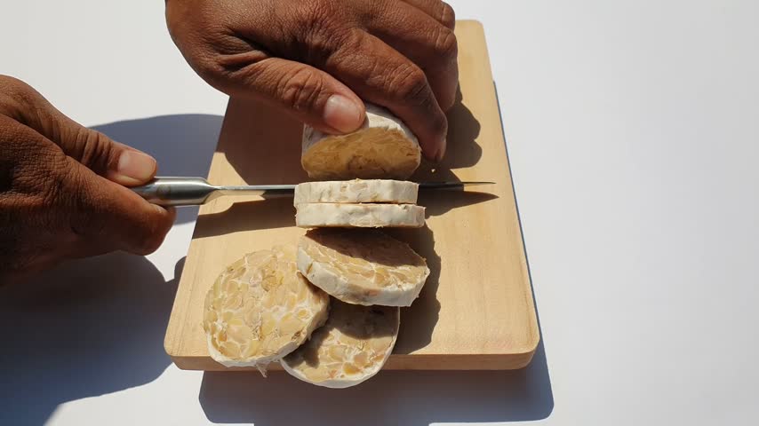 Close up of hands slicing raw tempeh or tempe on a wooden cutting board against a white background.