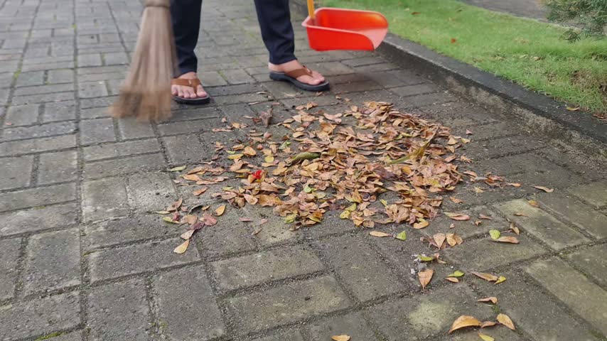 B-roll footage of a person sweeping a pile of dry autumn leaves on a concrete paving block pathway using a traditional stick broom and an orange plastic dustpan. Ideal for concepts of household chores, outdoor cleaning, autumn yard maintenance, and environmental care.