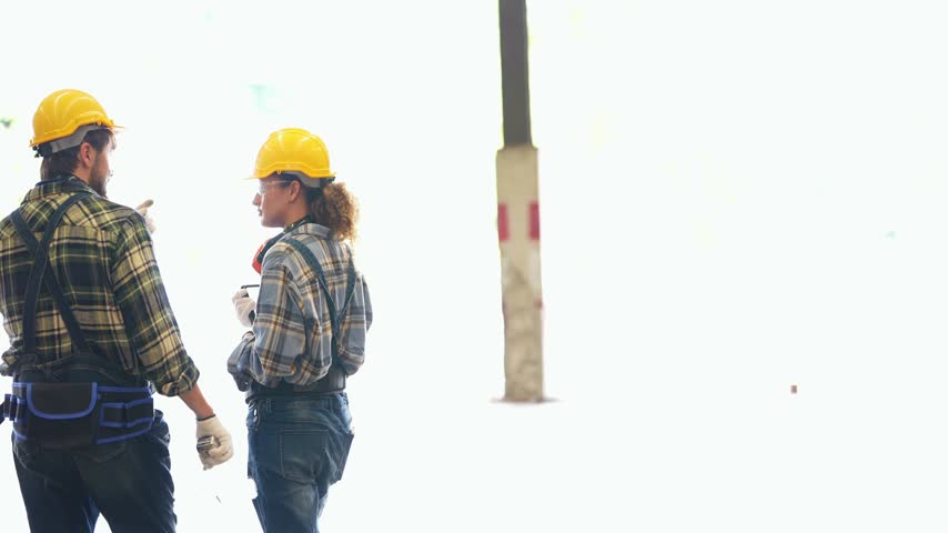 Wood factory: Bearded man and curly-haired woman in yellow hard hats walk out of bright daylight into a warehouse, the woman pausing to look around; real people.