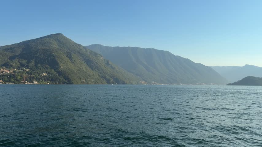 Wide scenic view of steep green mountains rising directly from a blue alpine lake, with small settlements visible along the shoreline in northern Italy.