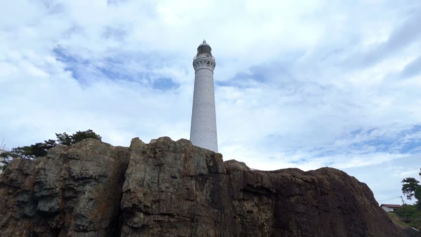 A majestic low-angle shot looking up at the historic Hinomisaki Lighthouse in Shimane, Japan. The striking white tower stands boldly on textured, rugged bedrock against a cloudy sky, providing a strong sense of scale.
