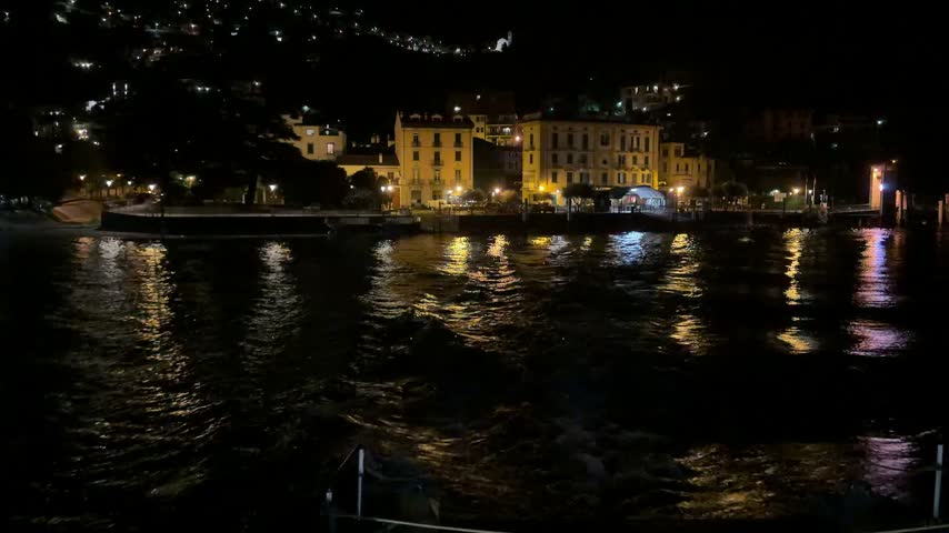 Atmospheric nighttime view of illuminated waterfront buildings reflecting across dark lake water, creating a peaceful European lakeside travel scene.
