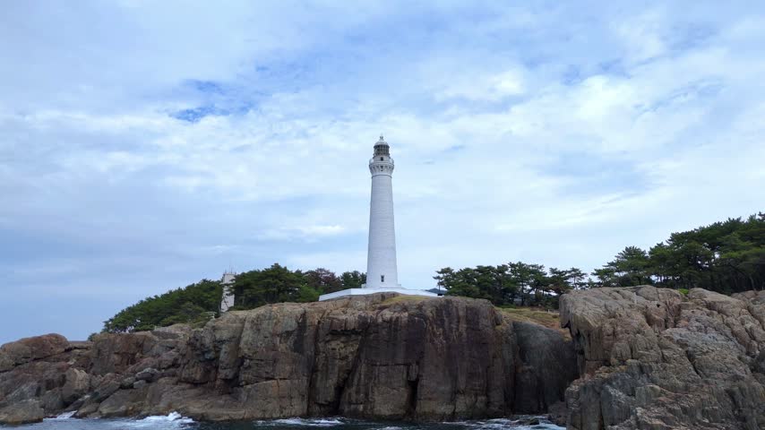 A beautiful ground-level shot looking up at the iconic white Hinomisaki Lighthouse standing on a rugged rocky cliff in Izumo, Shimane Prefecture, Japan. The majestic architecture offers a great establishing shot for travel documentaries.
