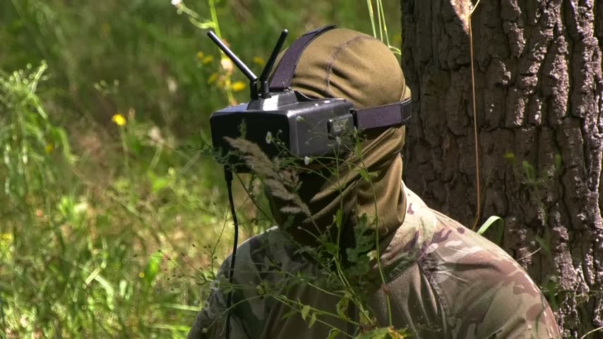 Military serviceman in uniform sitting in a forest clearing beside a tree and operating an FPV drone. Unmanned aerial technologies, drone control, military applications of UAVs, reconnaissance, and