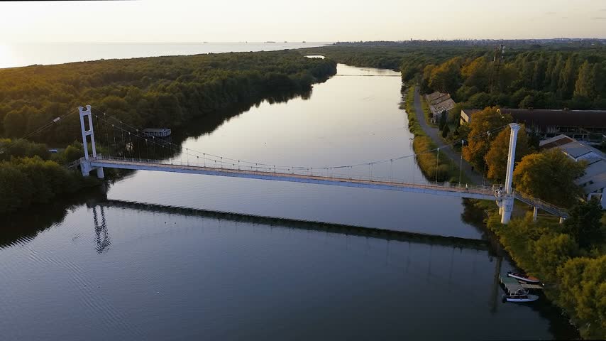 High-altitude drone shot capturing the iconic suspension pedestrian bridge over the river channel at Maltakva Park in Poti, Georgia. The video showcases lush green pine forest, calm river waters reflecting the sky, coastline of the Black Sea, and scenic autumn landscape at sunset. Perfect for travel, nature, aerial videography, and regional tourism projects.