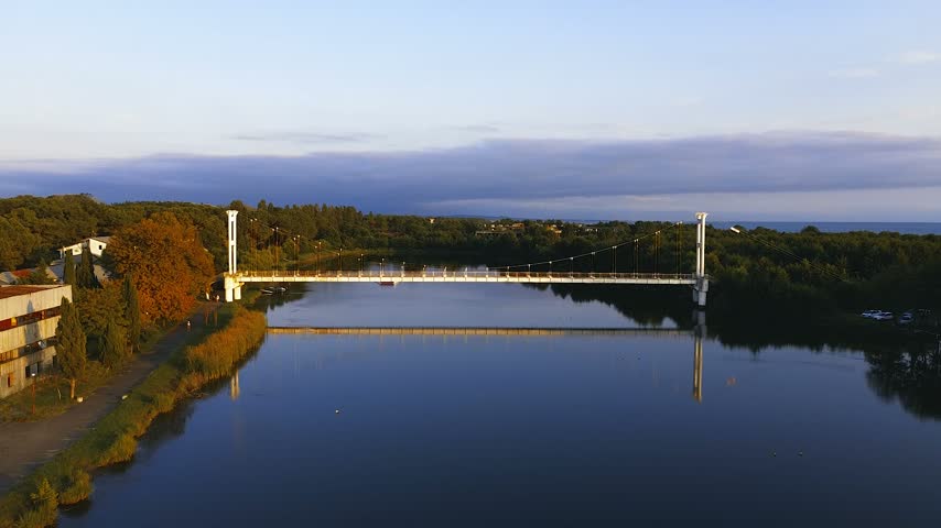 High-altitude drone shot capturing the iconic suspension pedestrian bridge over the river channel at Maltakva Park in Poti, Georgia. The video showcases lush green pine forest, calm river waters reflecting the sky, coastline of the Black Sea, and scenic autumn landscape at sunset. Perfect for travel, nature, aerial videography, and regional tourism projects.