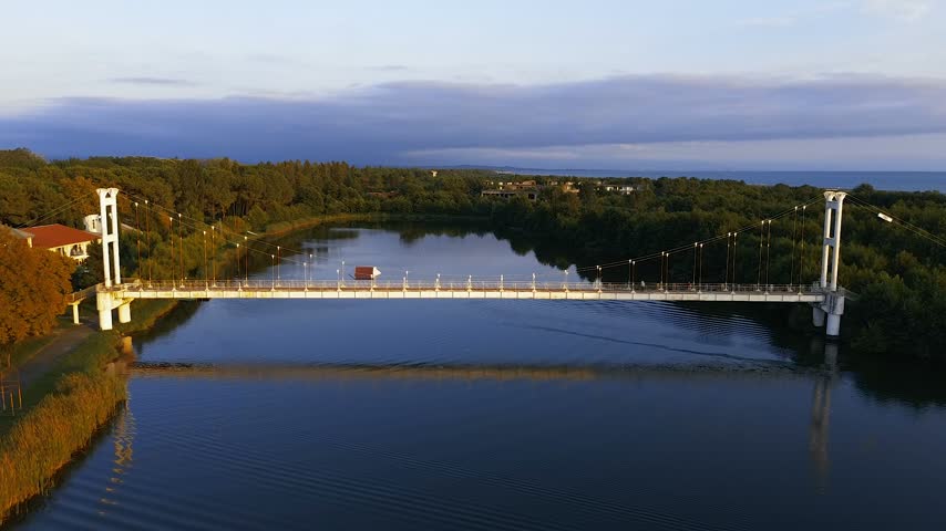 High-altitude drone shot capturing the iconic suspension pedestrian bridge over the river channel at Maltakva Park in Poti, Georgia. The video showcases lush green pine forest, calm river waters reflecting the sky, coastline of the Black Sea, and scenic autumn landscape at sunset. Perfect for travel, nature, aerial videography, and regional tourism projects.