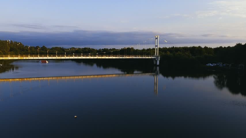 High-altitude drone shot capturing the iconic suspension pedestrian bridge over the river channel at Maltakva Park in Poti, Georgia. The video showcases lush green pine forest, calm river waters reflecting the sky, coastline of the Black Sea, and scenic autumn landscape at sunset. Perfect for travel, nature, aerial videography, and regional tourism projects.
