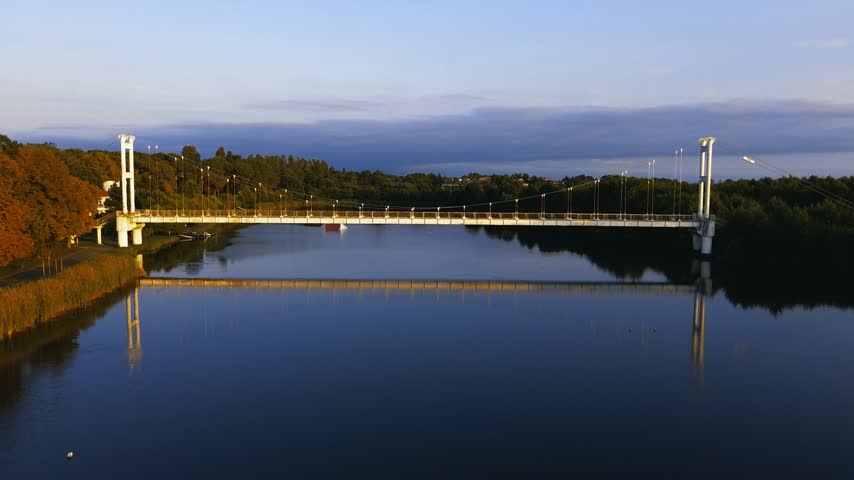 High-altitude drone shot capturing the iconic suspension pedestrian bridge over the river channel at Maltakva Park in Poti, Georgia. The video showcases lush green pine forest, calm river waters reflecting the sky, coastline of the Black Sea, and scenic autumn landscape at sunset. Perfect for travel, nature, aerial videography, and regional tourism projects.