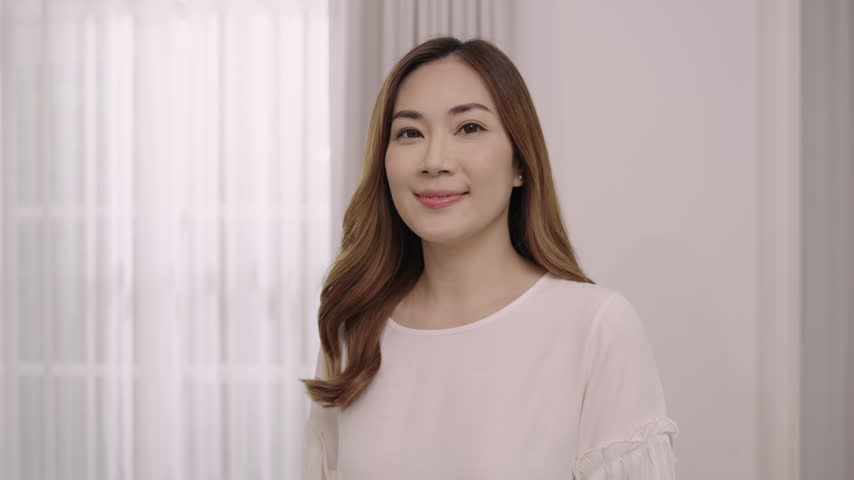 Portrait of happy and confident, beautiful Asian woman with long brown hair standing smiling while looking at the camera in the living room at home. Close-up attractive young female with Siam smile. 