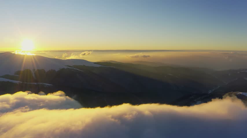 Cloud waves gently and slowly flow over the tops of snow-capped mountains covered with spruce forests at a cold sunset
