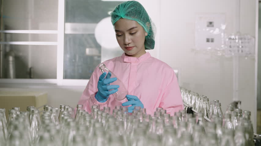 Happy beautiful Asian woman worker inspector in a factory uniform standing smiling and holding empty glasses beverage bottle while looking at camera on production line at the manufacturing industry.