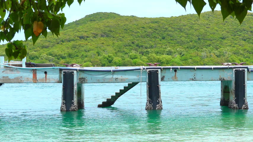 Wooden pier and ferry boat moored at tropical island harbor with clear blue ocean and green hills, clear sky