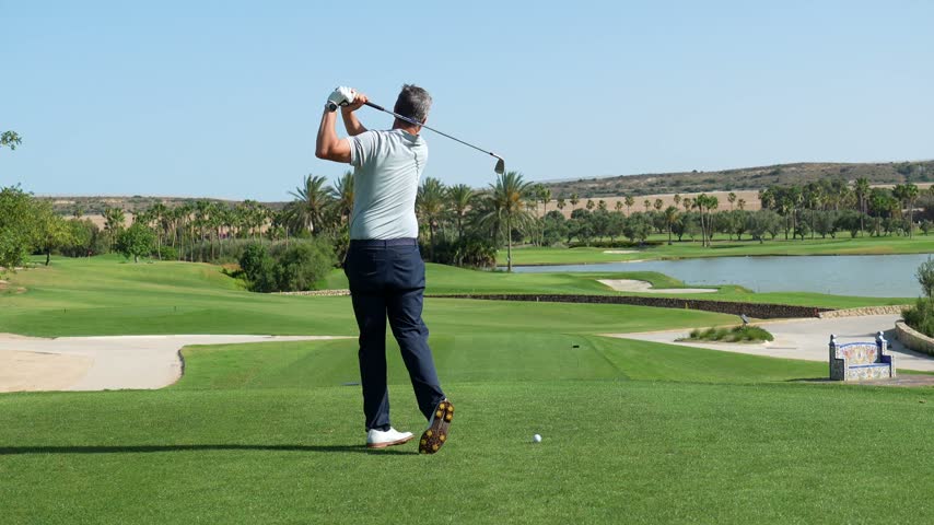 Professional golfer executing a powerful swing on a sunny day, driving the ball down the fairway of a luxury golf course with a lake and palm trees in the beautiful landscape