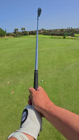 First person view of a golfer wearing a white glove, taking a few practice swings on a beautiful green fairway under a clear blue sky before hitting the ball on a sunny day