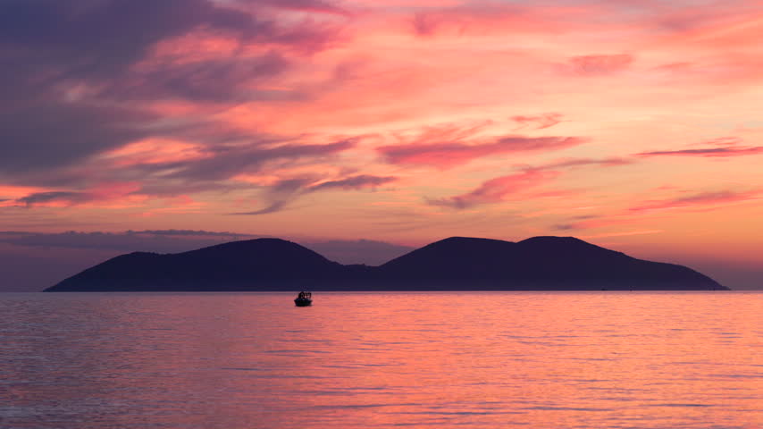 Stunning sunset over calm sea with distant islands silhouette and small boat floating on peaceful water surface during dusk. Adriatic sea near the city of Vlore in Albania, Europe.