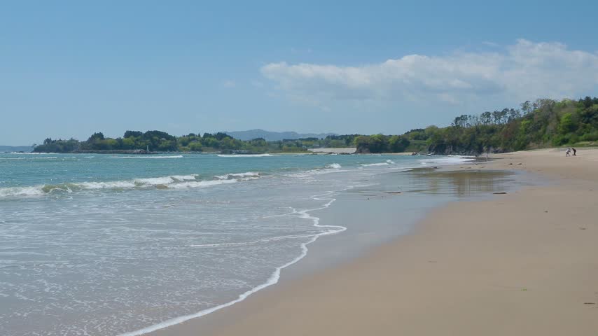 A smooth right-to-left sliding video showing a wide view of Otani Beach in Kesennuma, Miyagi Prefecture, Japan. The camera moves across the expansive sandy beach and calm blue sea under a clear sunny sky, capturing the beauty of the coastline.