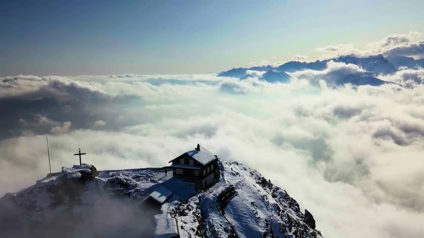 The image shows an aerial view of the Grosser Mythen mountain summit rising above a dense sea of clouds in Schwyz, Switzerland. The high-angle drone composition captures the rocky alpine peak piercing through an atmospheric cloud inversion layer under daytime conditions. This regional European geographical and natural asset is ideal for alpine travel documentaries, meteorological phenomenon features, and cinematic high-altitude aerial establishing sequences.