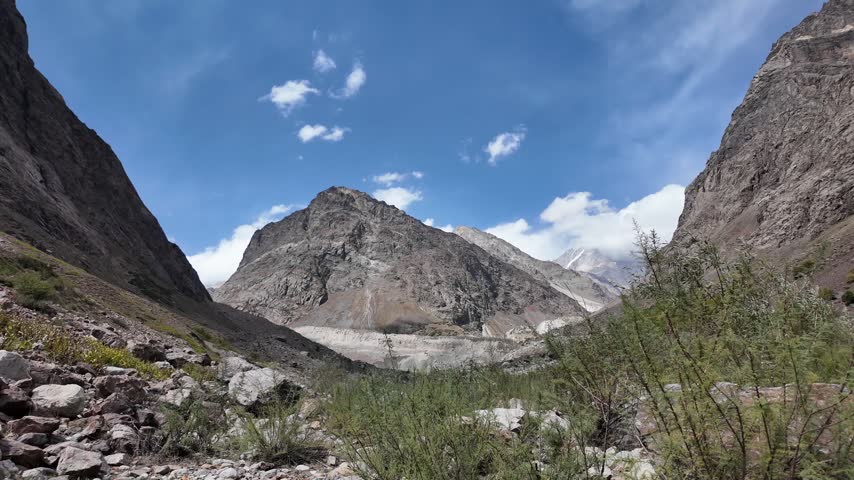 Remote high-altitude mountain valley with a rocky river flowing between rugged slopes and cloud-covered peaks in Northern Pakistan. Dramatic alpine wilderness landscape captured in 4K.