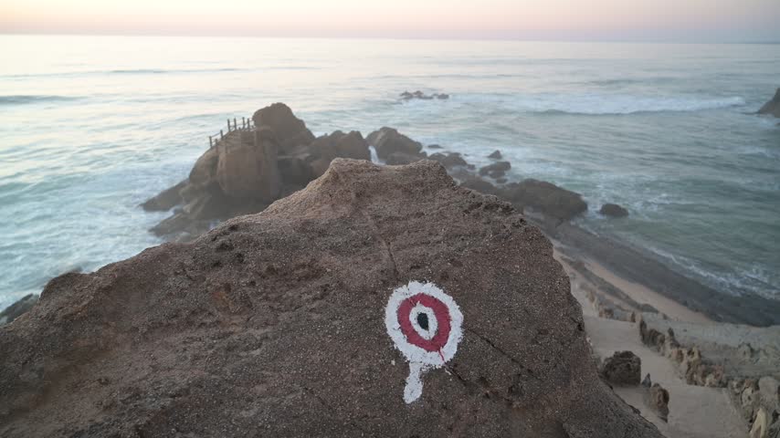 A painted trail marker on a rock overlooks waves and a rocky shore at Santa Cruz, Silveira, Portugal.
