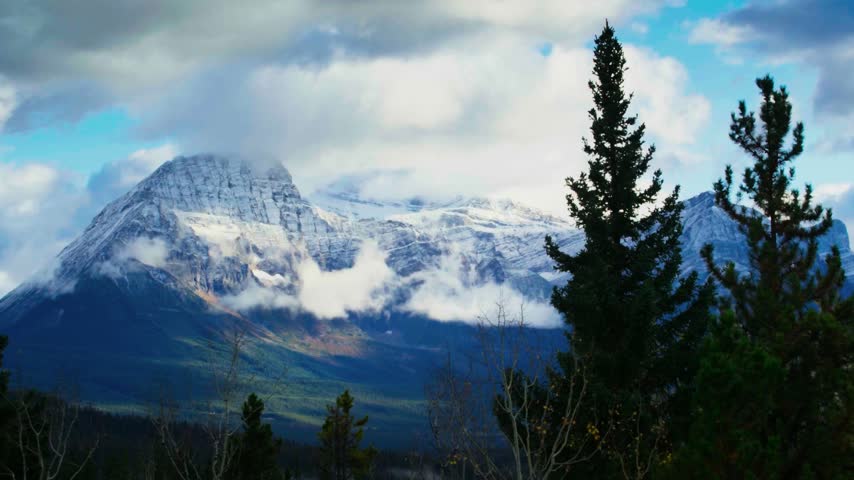 The image shows an aerial view of a snow-dusted mountain peak in Banff National Park, located in Alberta, Canada. The high-angle drone composition captures rugged rocky ridges, steep alpine slopes, and snow-covered terrain under daytime lighting conditions. This regional North American geographical and natural asset is ideal for Canadian travel documentaries, national park promotions, and cinematic high-altitude aerial establishing sequences.