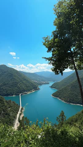Scenic view of turquoise Piva Lake winding between steep forest-covered mountains in Montenegro. A narrow mountain road follows the shoreline through the dramatic green landscape.