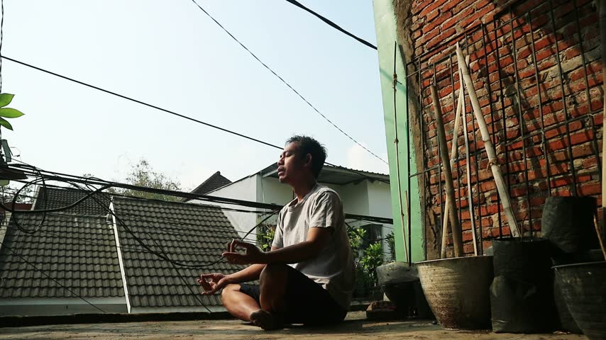 Indonesian man meditating outdoors in a relaxed pose, enjoying a peaceful moment in an urban setting.	