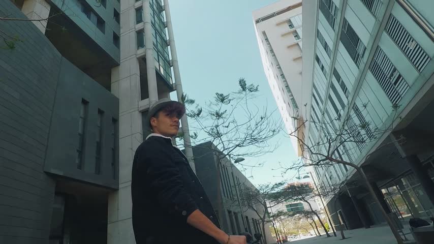 Cool young man with dyed hair and a hat walking confidently toward the camera on a city street, surrounded by tall modern architectural buildings under a clear blue sky