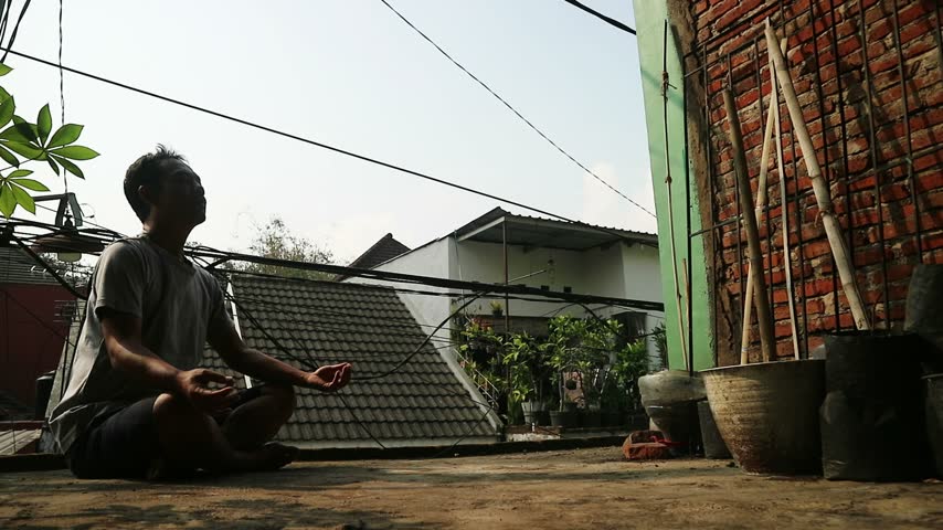 Indonesian man meditating outdoors in a relaxed pose, enjoying a peaceful moment in an urban setting.	