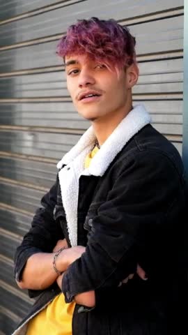 Cool young man with pink dyed hair and a lip piercing posing with arms crossed in front of a metal shutter, smiling and looking at the camera with a confident attitude