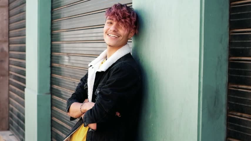 Happy young man with pink hair and a cool jacket leaning against a wall outdoors, smiling and looking at the camera with confidence while enjoying a sunny day in the city street