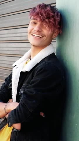 Cheerful young man with purple hair and piercings smiling at the camera. Handsome gen z male model posing outdoors with arms crossed against a wall in a city environment